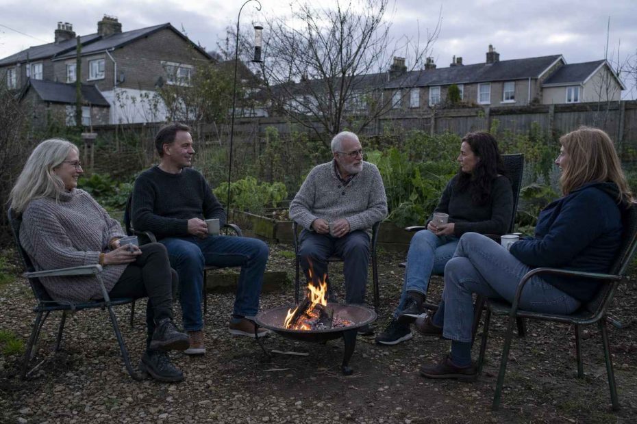 friends in jumpers around a simple metal fire pit on a gravel patch at dusk